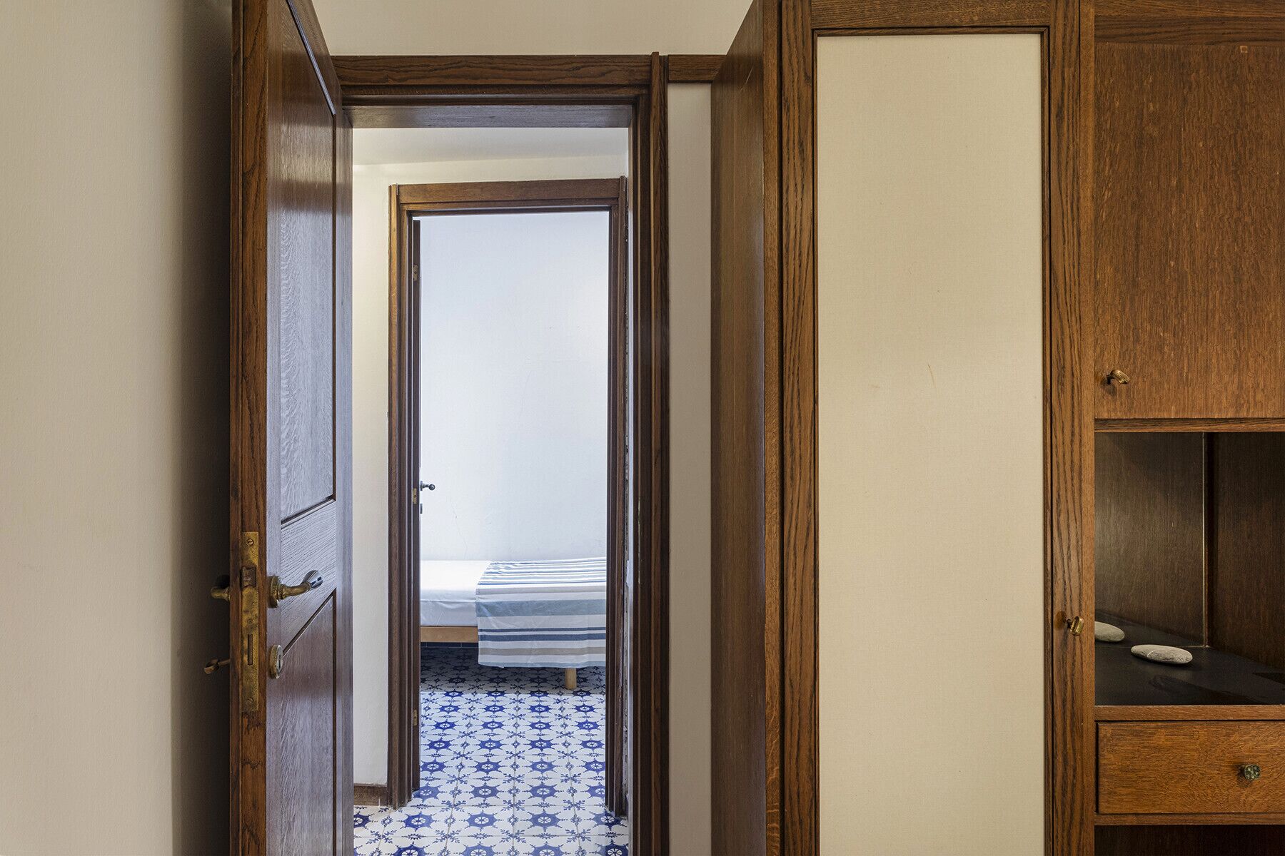 Un couloir étroit avec une porte en bois qui donne sur une pièce contenant un lit et un sol à motifs.