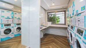 A modern laundry facility with multiple washing machines, a sink, and a window with a view of greenery. - Flat c Piscina e Academia na Ch Cara Klabin (São Paulo)