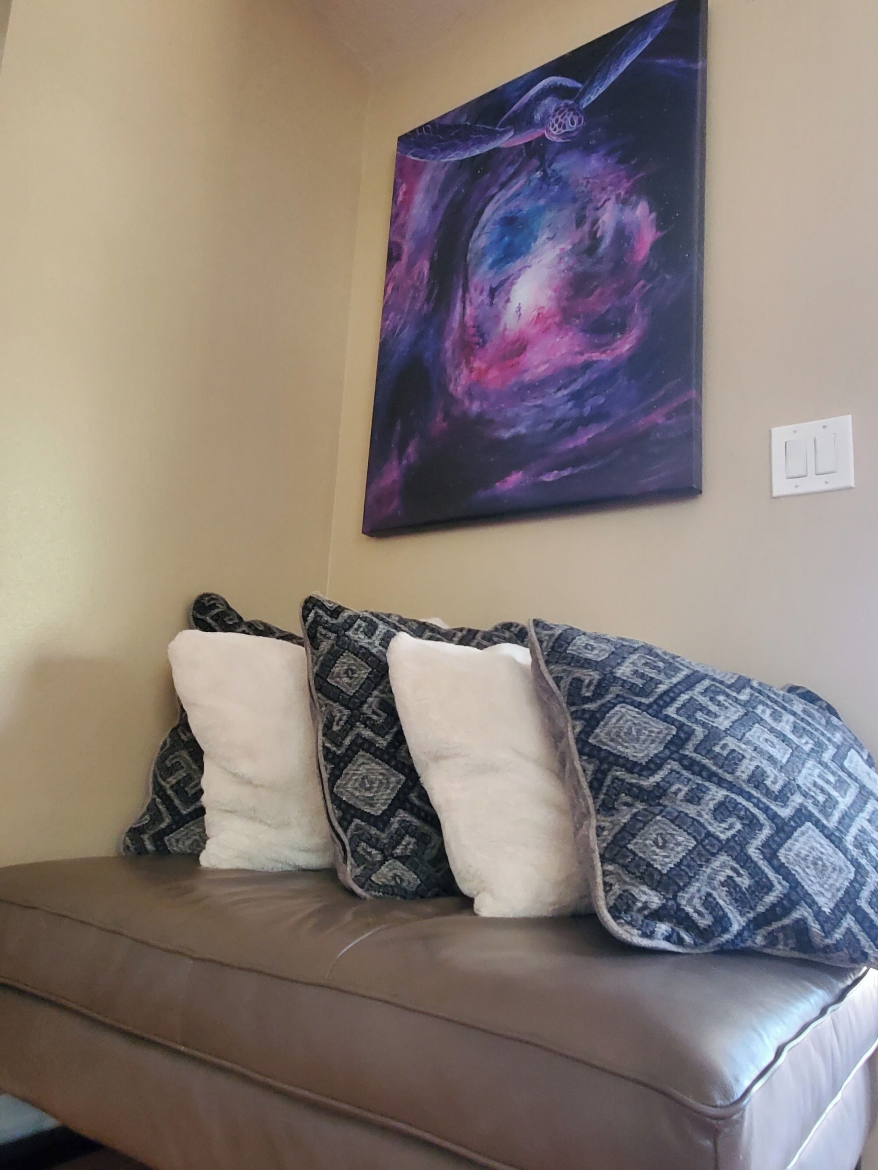 A brown leather sofa with patterned pillows against a beige wall, with a large abstract painting above.