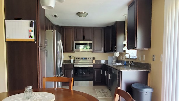 Une cuisine avec des meubles de rangement en bois foncé, des appareils électroménagers en acier inoxydable, une table à manger avec des chaises et une poubelle.