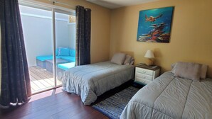 Une chambre avec deux lits, une table de chevet avec une lampe, un tableau représentant une tortue de mer et une porte-fenêtre coulissante donnant sur un espace extérieur aménagé pour se détendre.