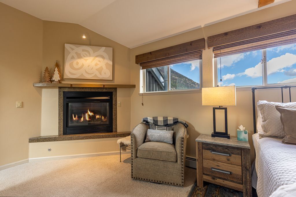 A cozy bedroom with a fireplace, a comfortable chair, a bedside table with a lamp, and a bed.