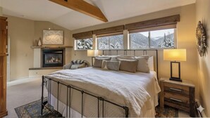 Une chambre à coucher avec un lit, une table de chevet, une lampe, une cheminée et des fenêtres donnant sur un paysage enneigé.