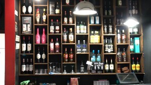 Zona de bar con estanterías llenas de botellas de alcohol, un cartel que dice 'CERVEZA ABIERTA AQUÍ' y una barra con una caja registradora.