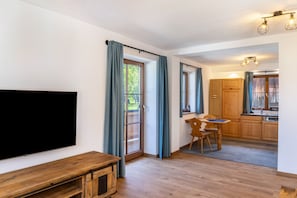 A modern living room with a flat-screen TV, wooden furniture, a dining area, and a kitchen with wooden cabinets.
