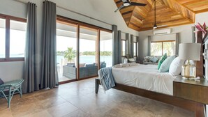 A spacious bedroom with a large bed, a ceiling fan, and a view of the ocean. - Enclave: Private Island, 7 Bungalows, All-Inclusive Luxury for Up to 22 Guests (Placencia)