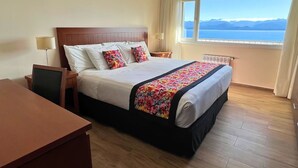 Et hotelværelse med seng, skrivebord, stol, natbord, lampe og et vindue med udsigt til bjerge og vand.