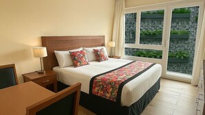 Habitación de hotel con cama, escritorio, silla, ventana con vistas a vegetación e iluminación en el techo.