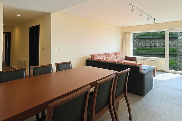 A modern living room with a sofa, a dining table, and chairs. There is a large window with a view of greenery outside.