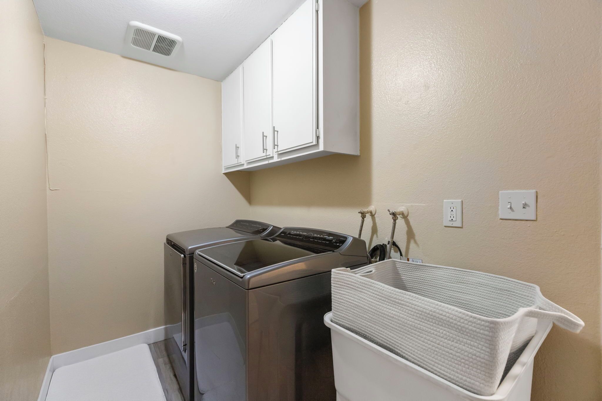 A compact laundry room with a front-loading washing machine, a white cabinet above, and a white laundry basket on the floor.