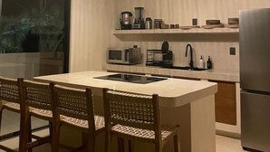 A modern kitchen with a central island, two wicker chairs, a stainless steel refrigerator, and pendant lighting. - Casa Hood Boutique Hotel (Isla Holbox)
