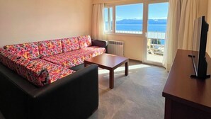 A room with a floral sofa, a wooden coffee table, a TV on a stand, and a large window with a view of the sea and mountains.