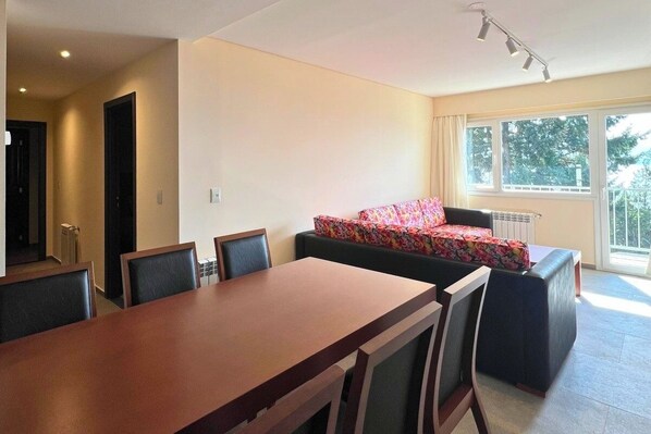 A dining area with a long table and chairs, a sofa with a floral cover, and a window with a view of trees.