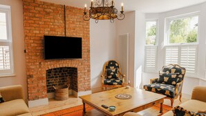 Un salon doté d’une cheminée en briques, d’un téléviseur à écran plat fixé au-dessus, de deux fauteuils à motifs, d’une table basse en bois et d’un lustre.