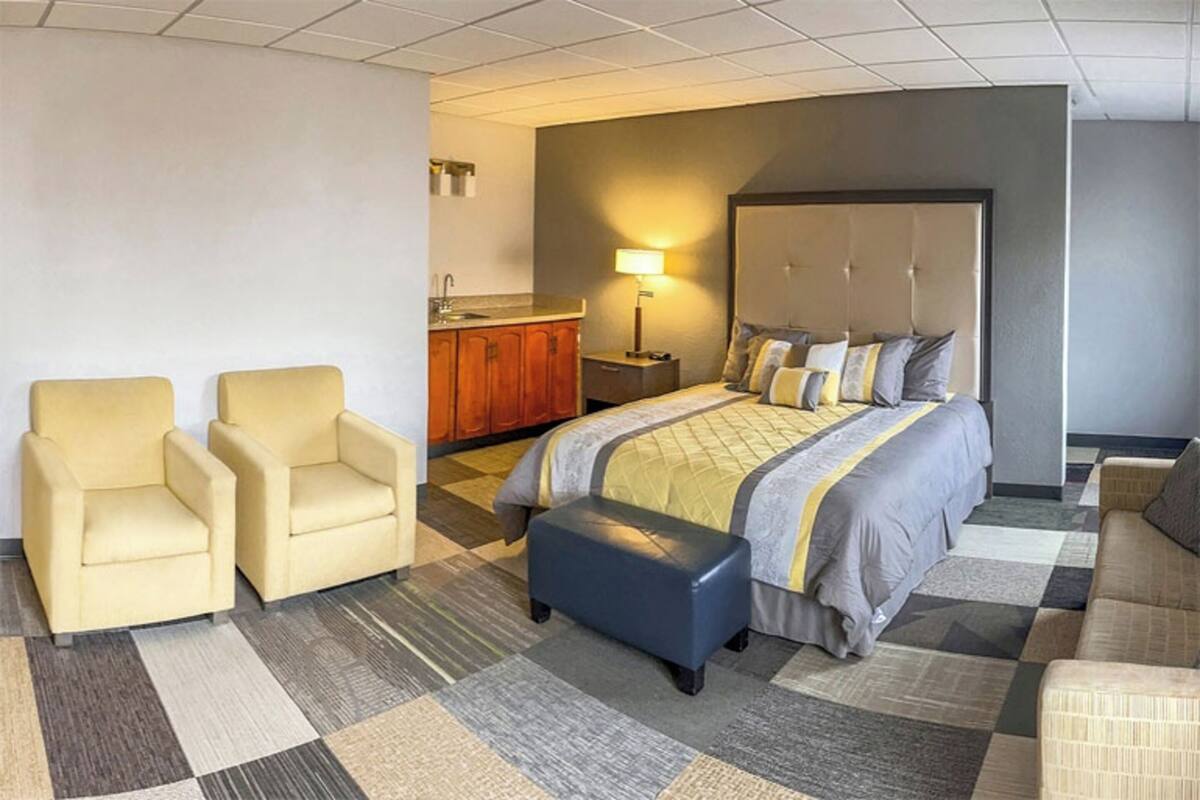 A hotel room with a bed, two armchairs, a bench, a nightstand, a lamp, and a small kitchenette.