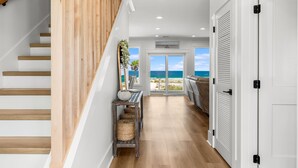 A modern interior with a wooden staircase, a console table with baskets, and a view of the ocean through large doors. - Luxury Gulf-front Retreat | Sleeps 24 | Private Pool (Port St. Joe)