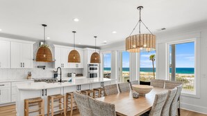 A modern kitchen with white cabinetry, a large island, and a dining area with wicker chairs and a view of the beach. - Luxury Gulf-front Retreat | Sleeps 24 | Private Pool (Port St. Joe)