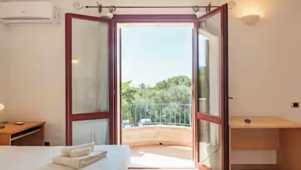 A hotel room with a bed, a desk, and a balcony with a view of greenery.