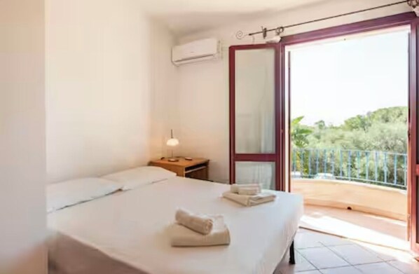 A bedroom with a bed, a nightstand, a lamp, and a balcony with a view of greenery.