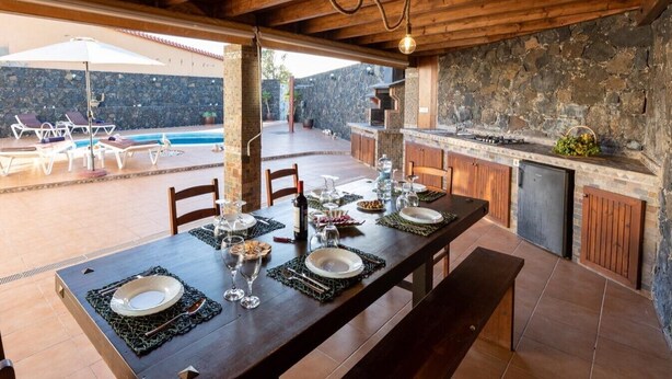 An outdoor dining area with a stone wall, wooden ceiling, and a pool in the background. - Private Paradise in Fuerteventura · Pool, Barbecue, and Dream Beaches (CN)