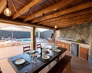 Un espacio de comedor al aire libre con pared de piedra, techo de madera y una piscina al fondo.