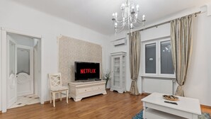 Een woonkamer met een televisie, witte meubels, een kroonluchter en een raam met gordijnen.