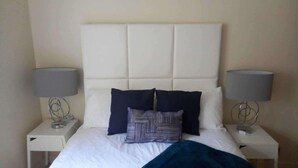 A neatly made bed with a white headboard, two dark pillows, and a blue blanket. There are two bedside tables with lamps on either side of the bed. - Gourock Blissful Apartment With 3 Bedrooms (Gourock)