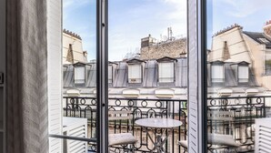 Un balcon avec une table et une chaise blanches, une rambarde en métal et une vue sur les bâtiments voisins.
