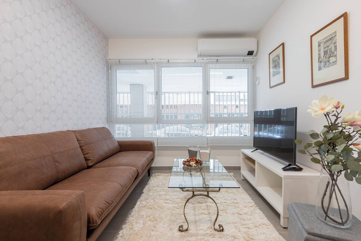 A living room with a brown sofa, a glass coffee table, a TV on a stand, and a vase with flowers.