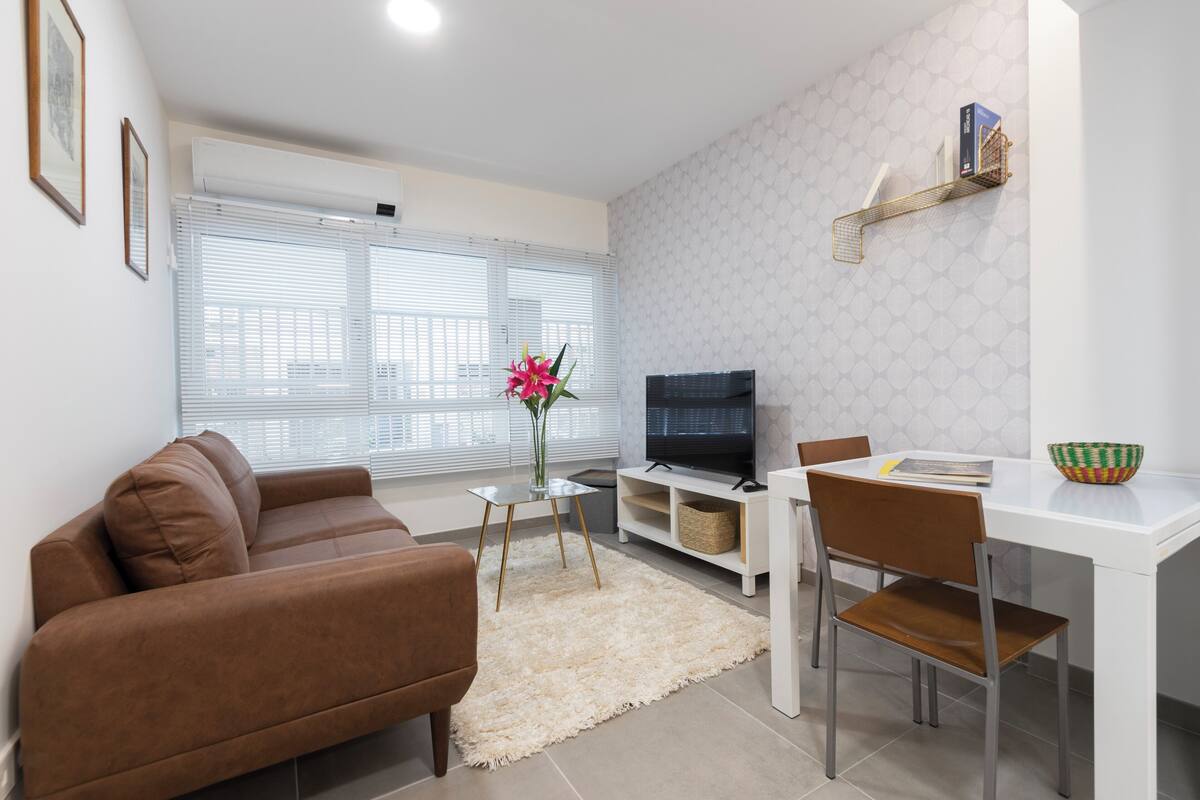 A modern living room with a brown sofa, a TV, a dining table with a chair, and a decorative vase.