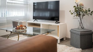 A modern living room with a flat-screen TV on a white stand, a glass coffee table with decorative items, a grey ottoman, and a vase with flowers.