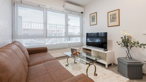 A living room with a brown sofa, a glass coffee table, a flat-screen TV on a white stand, and a vase with flowers on a grey side table.