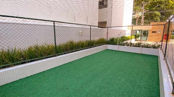 A fenced outdoor area with artificial green turf, surrounded by a white tiled wall and a chain-link fence. - Apto c Varanda Piscina e Sal o de Jogos (Salvador)