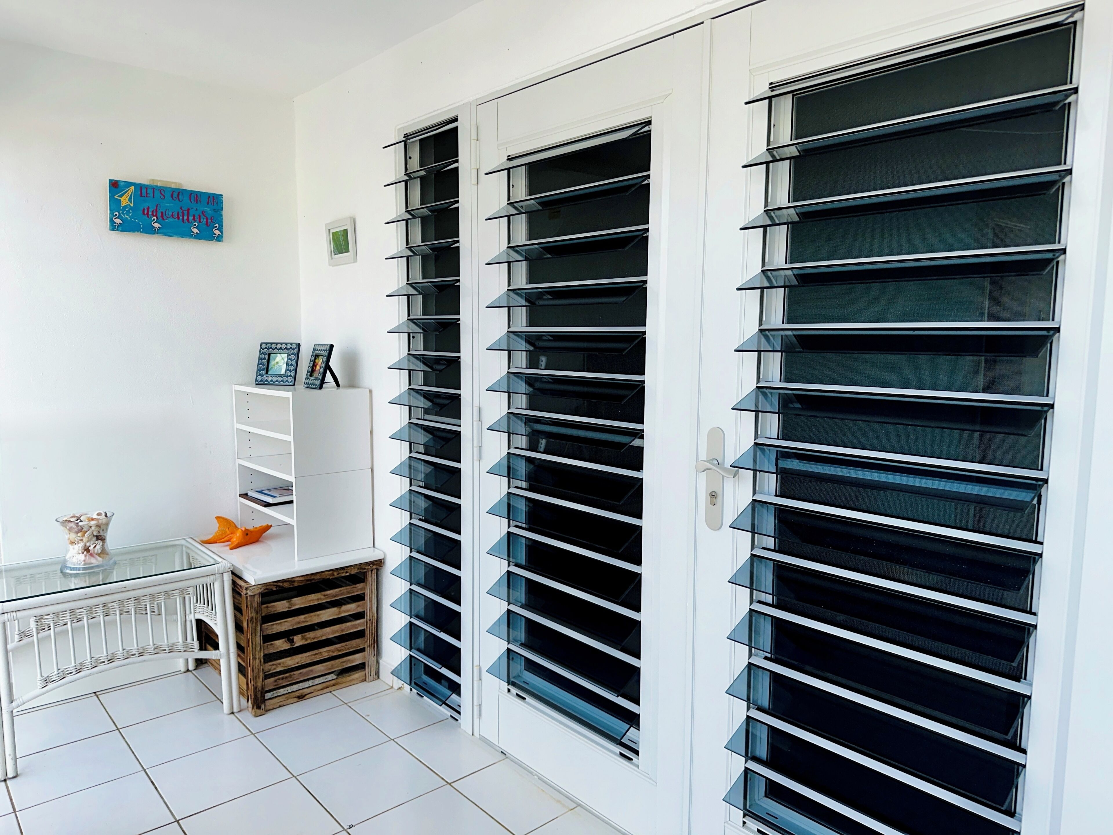 A room with a white door, a white shelf with framed pictures, a glass table, and a wooden crate.