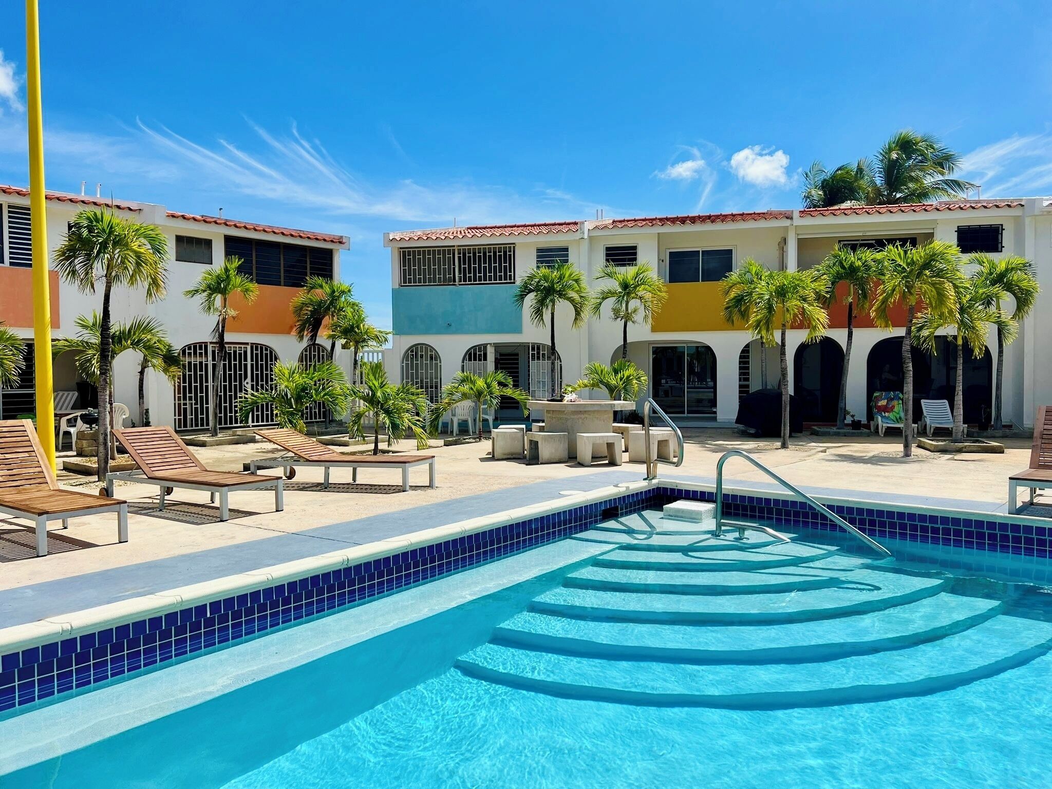 A pool area with lounge chairs, palm trees, and a building with a tiled roof.