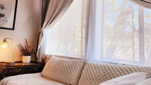 Ein Schlafzimmer mit Holzboden, einem Bett mit weißer Bettwäsche, einem Nachttisch mit Lampe und einem Fenster mit durchscheinenden Vorhängen.