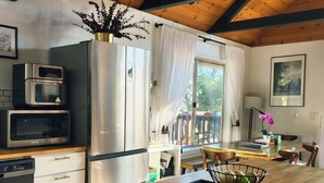 Eine moderne Küche mit einem Edelstahlkühlschrank, einer Mikrowelle und einem Geschirrspüler. Ein Holztisch mit Stühlen, ein Fenster mit Vorhängen und ein kleiner Balkon mit Blick auf Bäume.