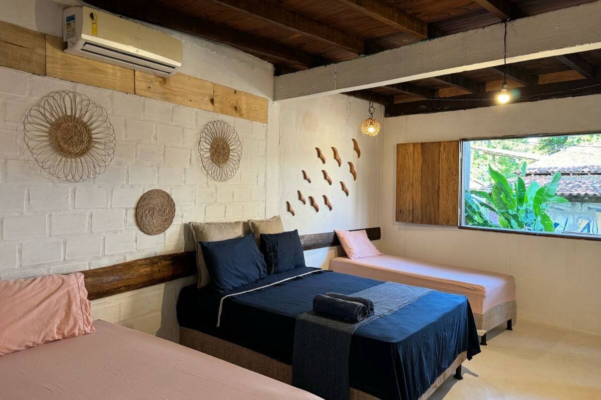A bedroom with two beds, a white brick wall, wooden beams, and a window with a view of greenery.