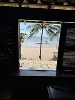 Vista da praia através de uma janela, com palmeira, praia e barcos.