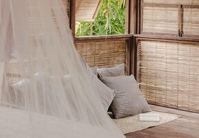 Quarto com cama coberta por mosquiteiro, paredes de madeira e janela com vista para vegetação.