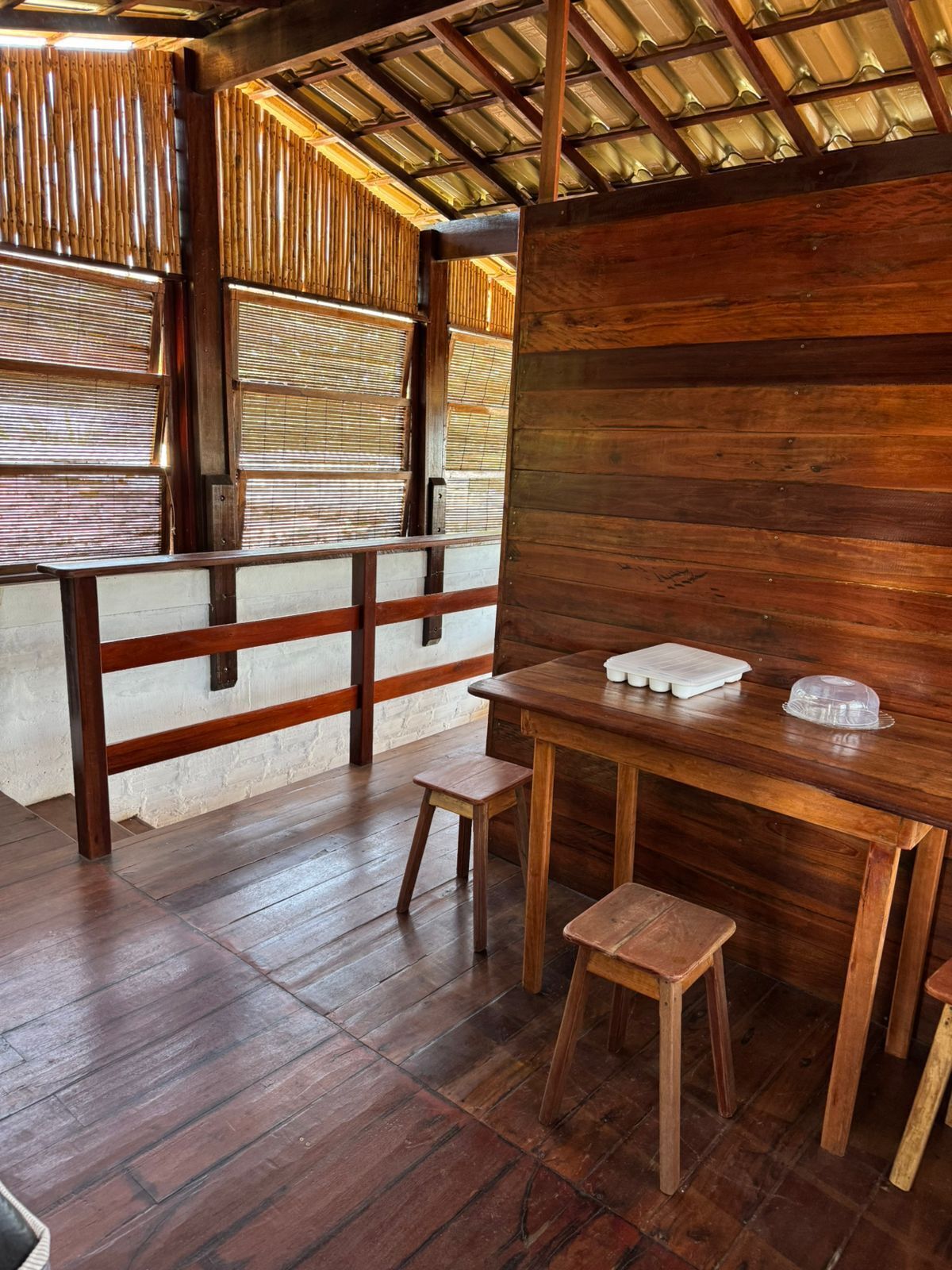 Un interior de madera con una mesa y taburetes, un muro de bambú y un techo de metal.
