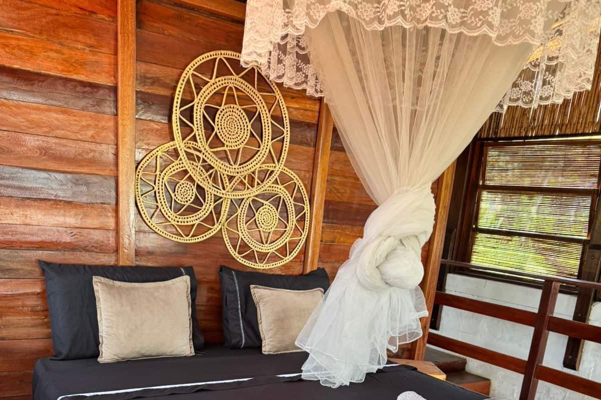 A double bed with a grey bedspread, a canopy, and rolled towels. There are three pillows on the bed, and a woven wall decoration above it.