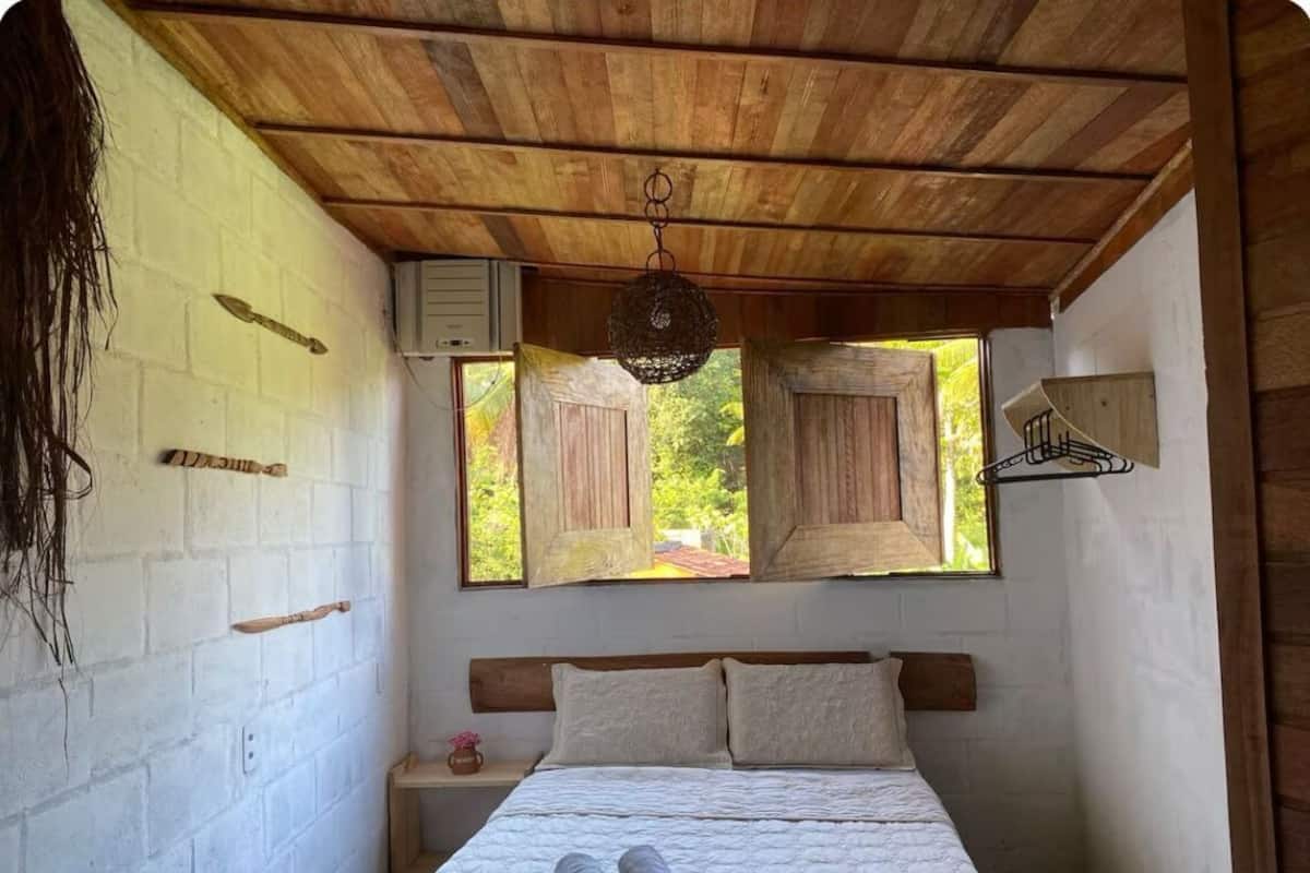 A bedroom with a wooden ceiling, a bed with two pillows, a nightstand, and a window with shutters.