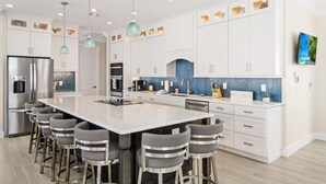 A modern kitchen with white cabinets, a large island with bar stools, stainless steel appliances, and pendant lighting. - Anchors Down (Fort Myers Beach)