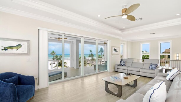 A spacious living room with a large sliding glass door leading to a balcony with a view of the ocean. There is a grey sofa, a blue armchair, and a coffee table with a book and a bowl. - Anchors Down (Fort Myers Beach)