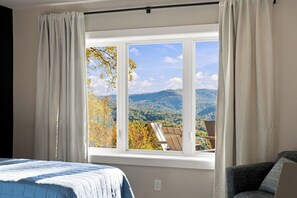 A bedroom with a large window, a bed, a chair, and a view of a mountainous landscape.