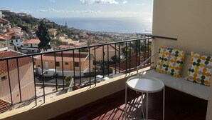 En balkon med et hvidt bord og en sofa med prikkede puder, med udsigt over en kystby med terracotta-tage og havudsigt.