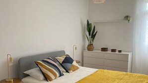 Et moderne soveværelse med en stor seng, et sengebord med lampe, en hængende lysarmatur og en potteplante på en kommode.