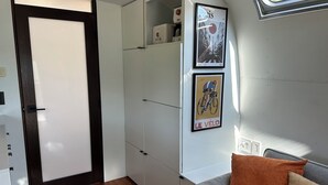 En kompakt opholdsstue med dør, hvide skabe, en grå sofa med orange puder og to billeder i rammer på væggen.