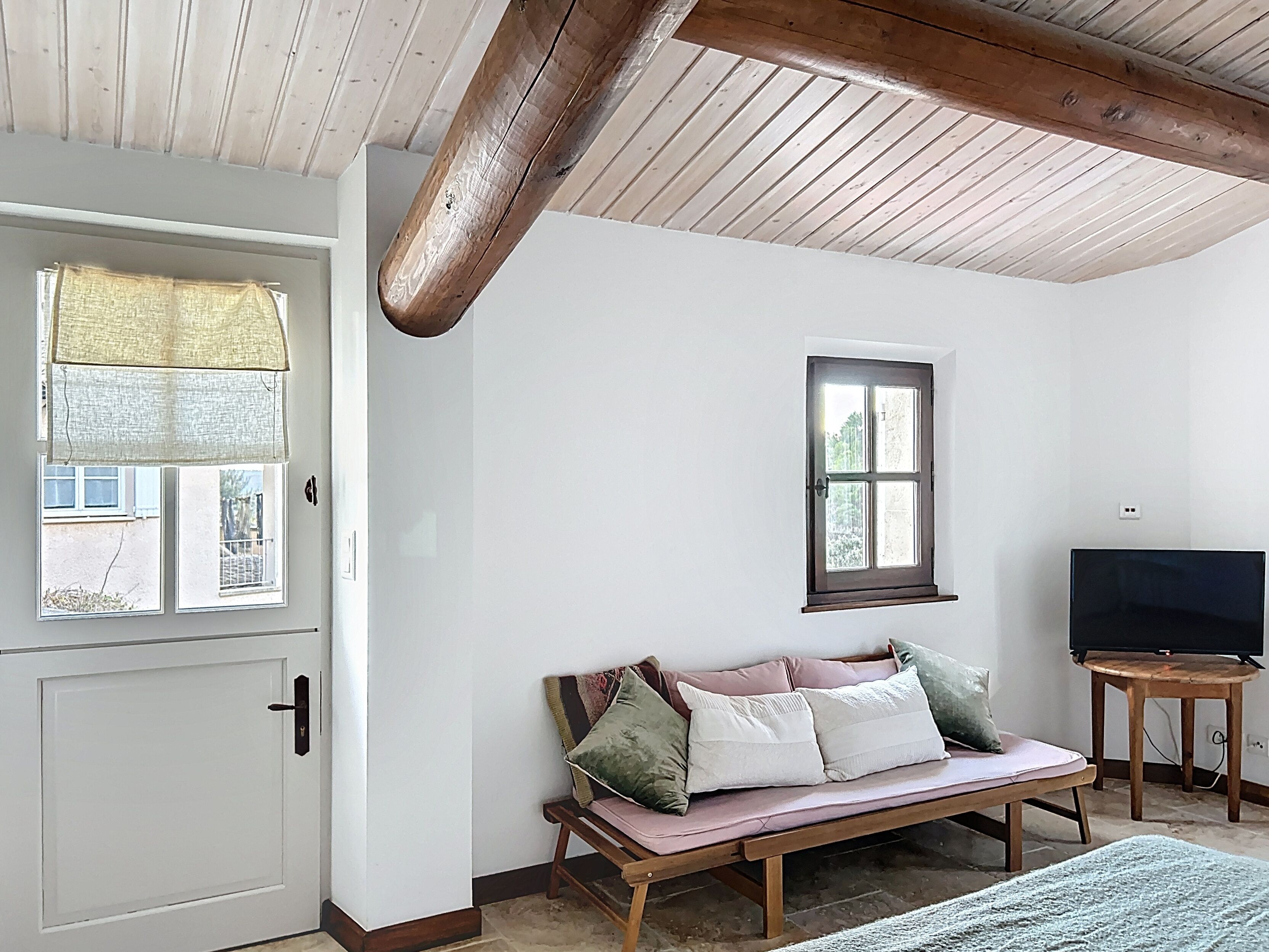 Une pièce avec un plafond en bois, une porte blanche, une fenêtre avec un rideau, un banc en bois avec des coussins et un téléviseur posé sur une petite table.